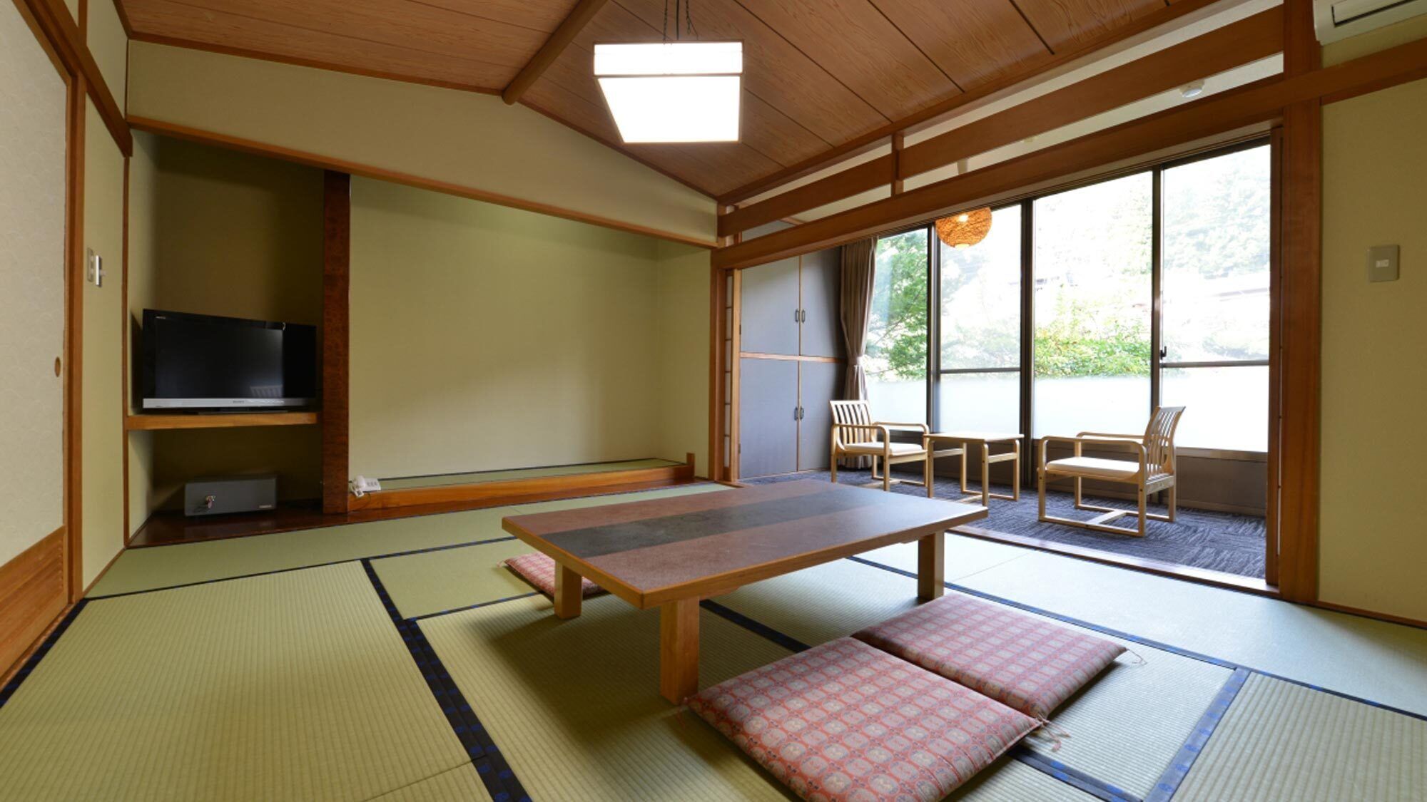 Una habitación japonesa tradicional con suelo de tatami, una mesa baja, cojines, un televisor y puertas correderas que dan a un área exterior.