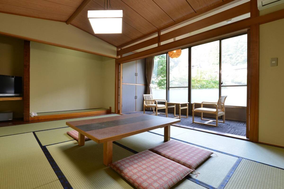 Um quarto japonês tradicional com tatames, uma mesa baixa, almofadas, televisão e portas deslizantes que dão para uma área externa.