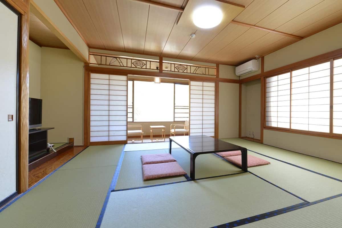 Um quarto japonês tradicional com tatames, uma mesa baixa, almofadas, portas deslizantes e uma televisão.