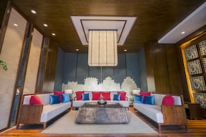 A modern living room with a wooden ceiling, a central coffee table, and a sofa set with red and blue cushions.