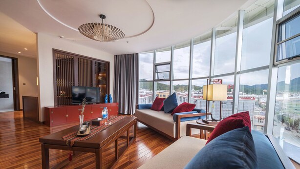 Ein modernes Wohnzimmer mit einer Couch, einem Couchtisch und einem Fernseher auf einer Holzkommode. Große Fenster bieten Blick auf die Stadt.