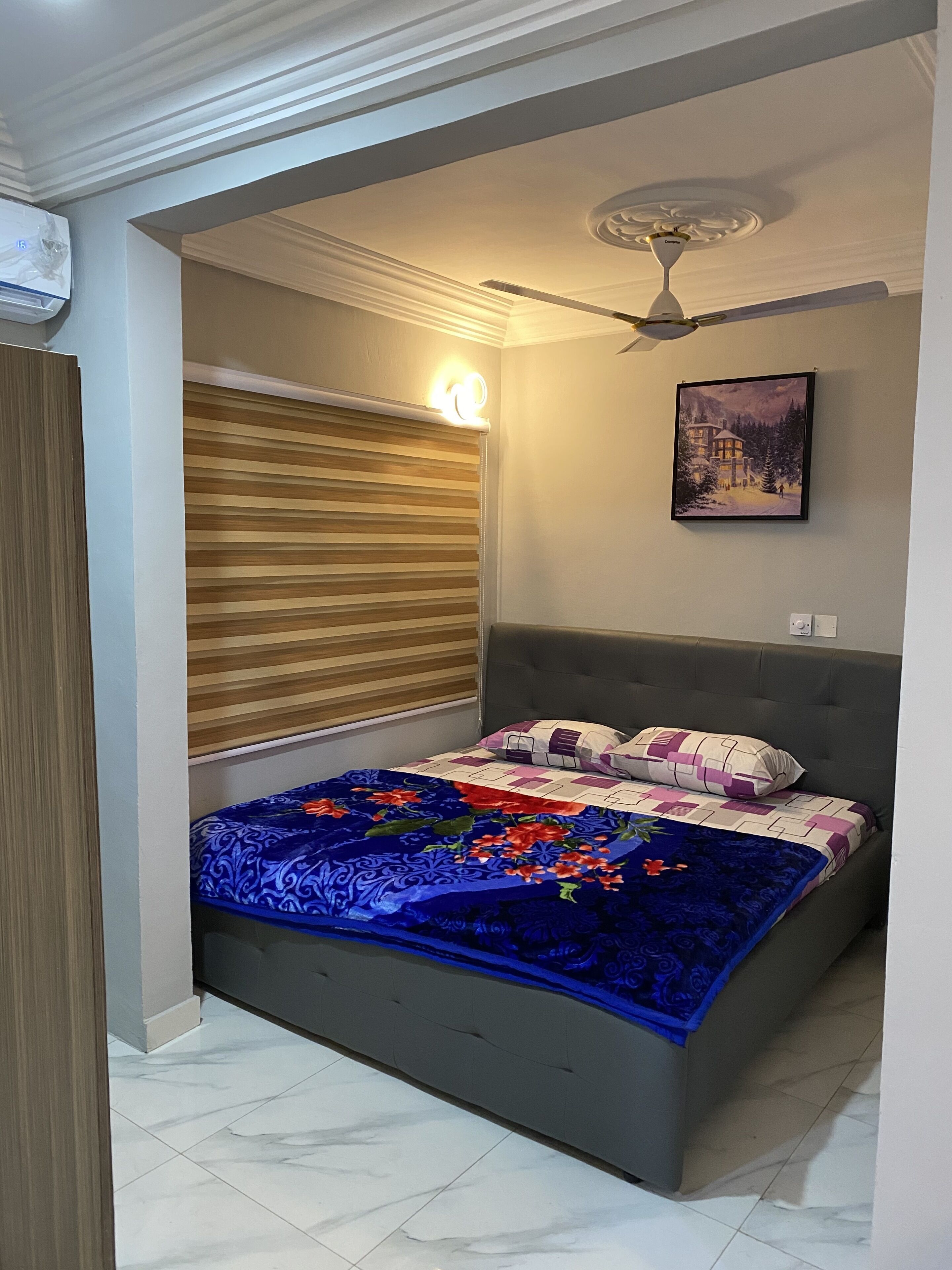 A bedroom with a bed, a ceiling fan, a wall-mounted light, and a framed picture.