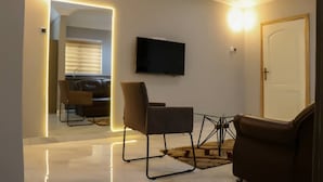 Una sala de estar moderna con un televisor de pantalla plana montado en la pared, dos sillones de cuero, una mesa de centro de vidrio y un espejo que refleja la habitación.