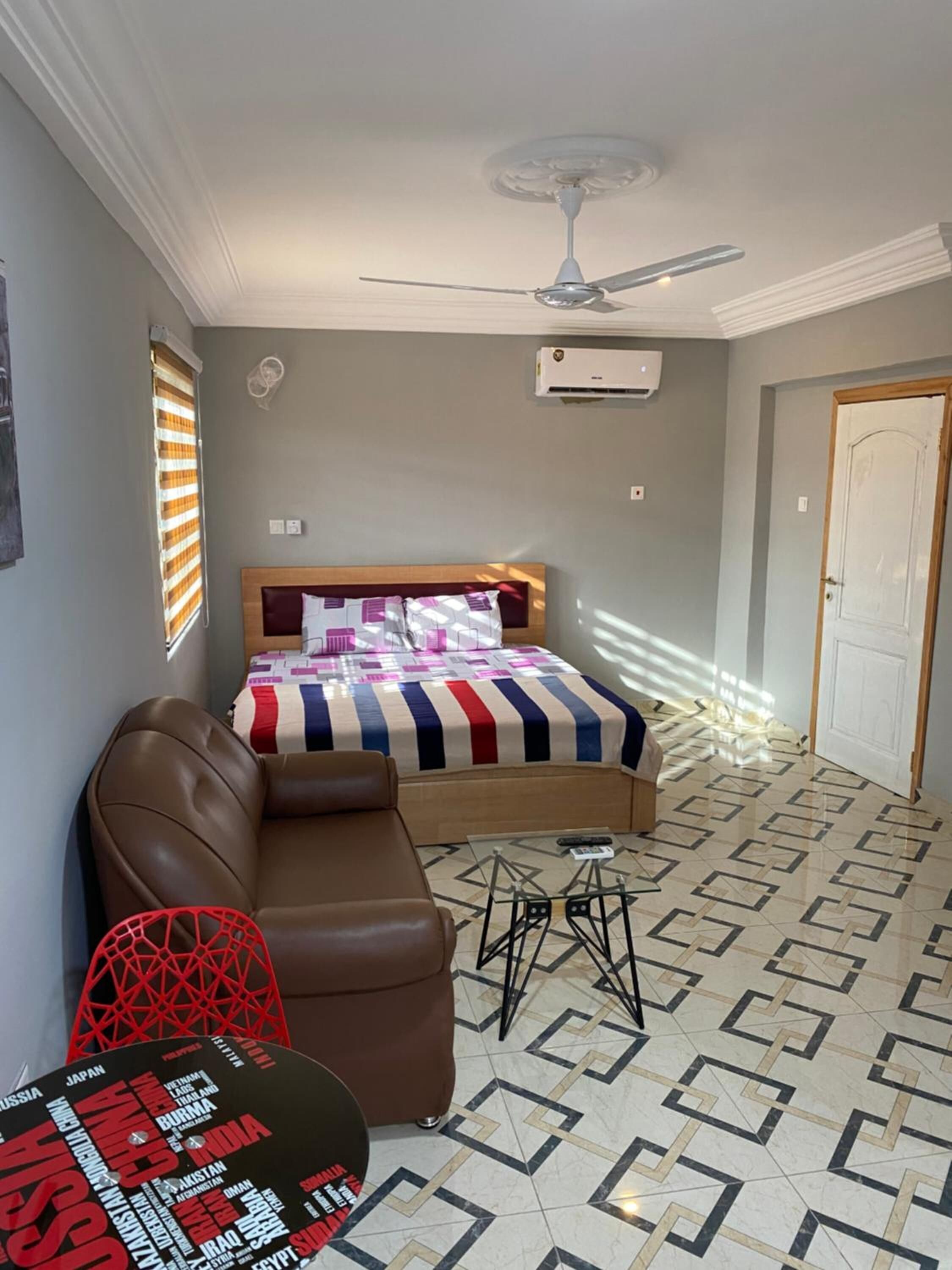 Ein Schlafzimmer mit Bett, Deckenventilator, Klimaanlage, Fenster mit Jalousien, Tür und einem gemusterten Boden.