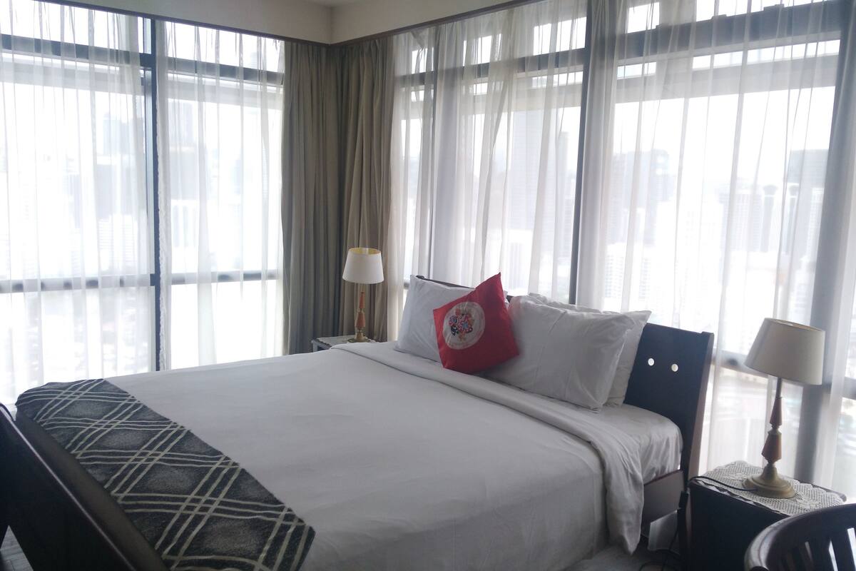 A hotel room with a large bed, two bedside tables with lamps, a chair, and a view of the city through sheer curtains.
