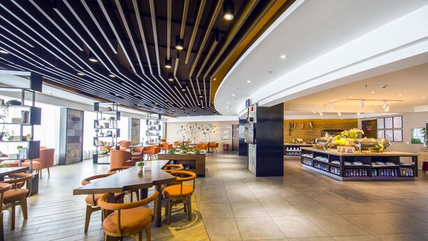 A spacious restaurant with wooden tables and orange chairs, a curved ceiling with linear lighting, and a central display area with various items. - Yunman Hotel Kunming Donghua (Kunming)