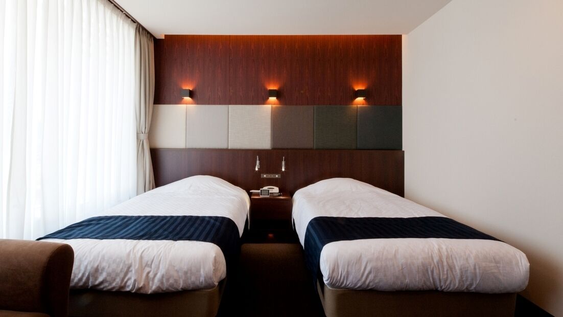 A hotel room with two single beds, a wooden headboard, wall-mounted lights, and a nightstand.