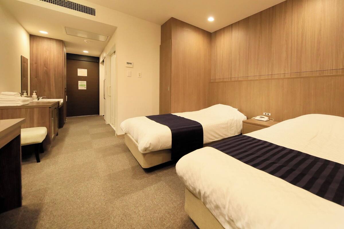Ein Hotelzimmer mit zwei Betten, einem Schreibtisch mit Stuhl und einem durchgegangenen Türspalte sichtbaren Badezimmer.