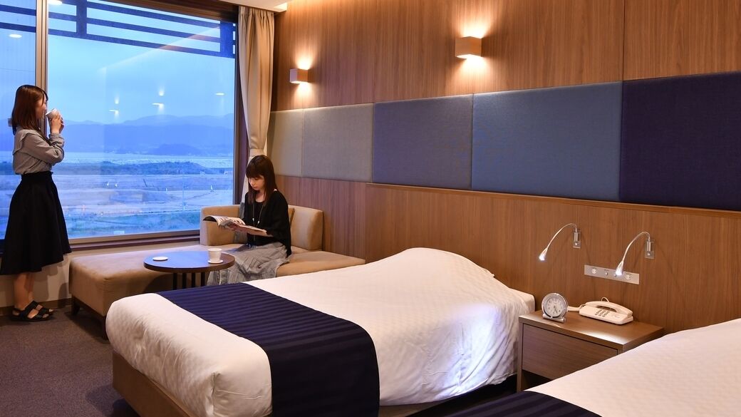 A hotel room with two beds, a small table, a chair, a bedside table with a phone and alarm clock, and a window with a view of the landscape.