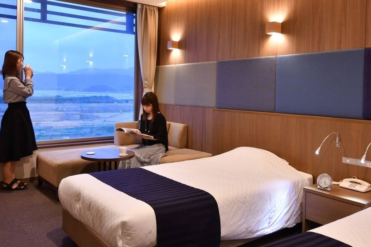 Ein Hotelzimmer mit zwei Betten, einem kleinen Tisch, einem Stuhl, einem Nachttisch mit Telefon und Wecker sowie einem Fenster mit Blick auf die Landschaft.