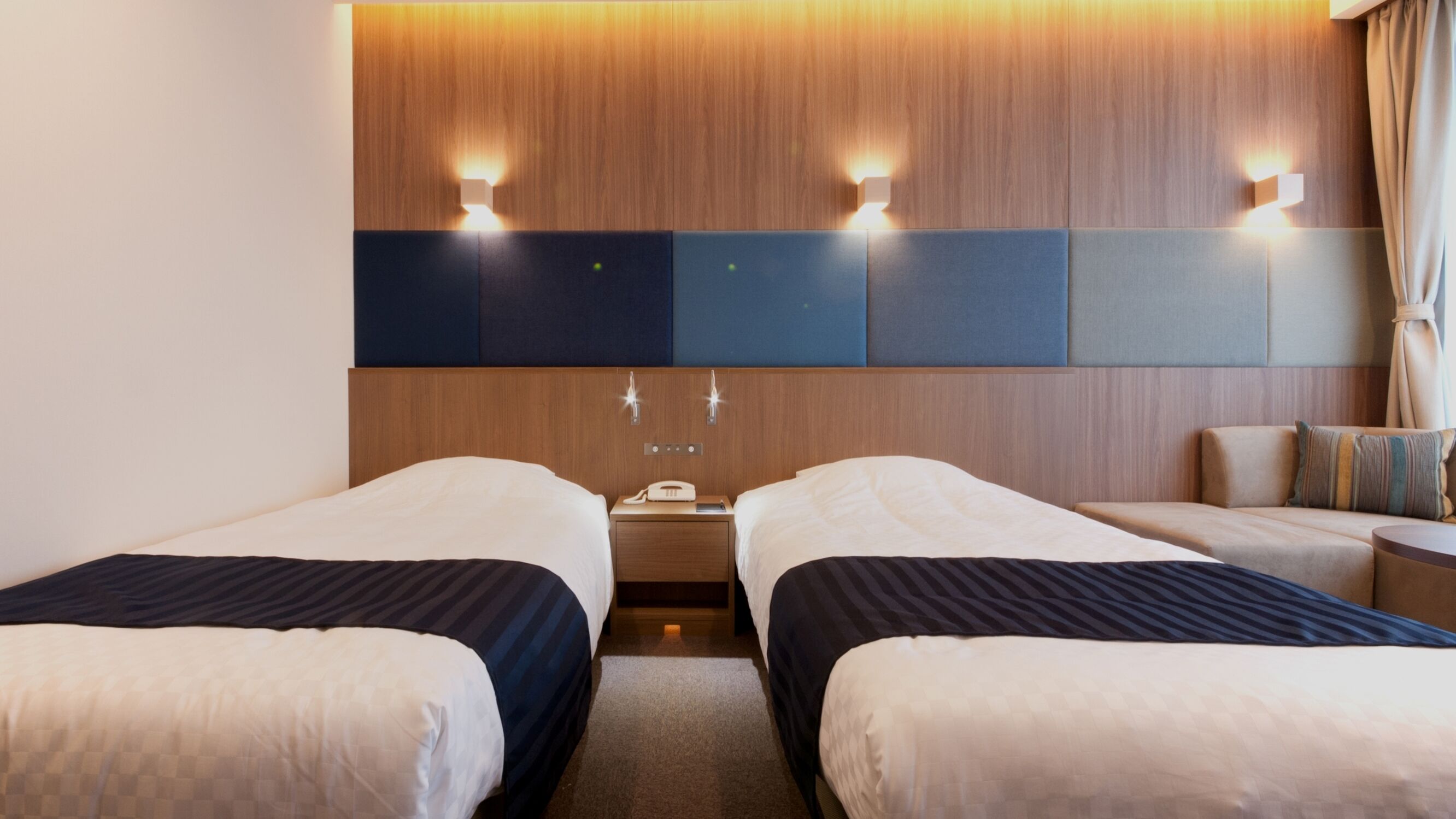 A hotel room with two beds, a wooden headboard, a nightstand with a phone and lamp, and a seating area with a sofa and armchair.