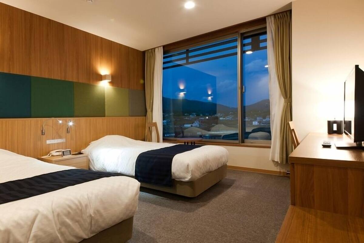 Ein Hotelzimmer mit zwei Betten, einem Schreibtisch, einem Stuhl, einem Fernseher und einem großen Fenster mit Blick auf die Stadt.