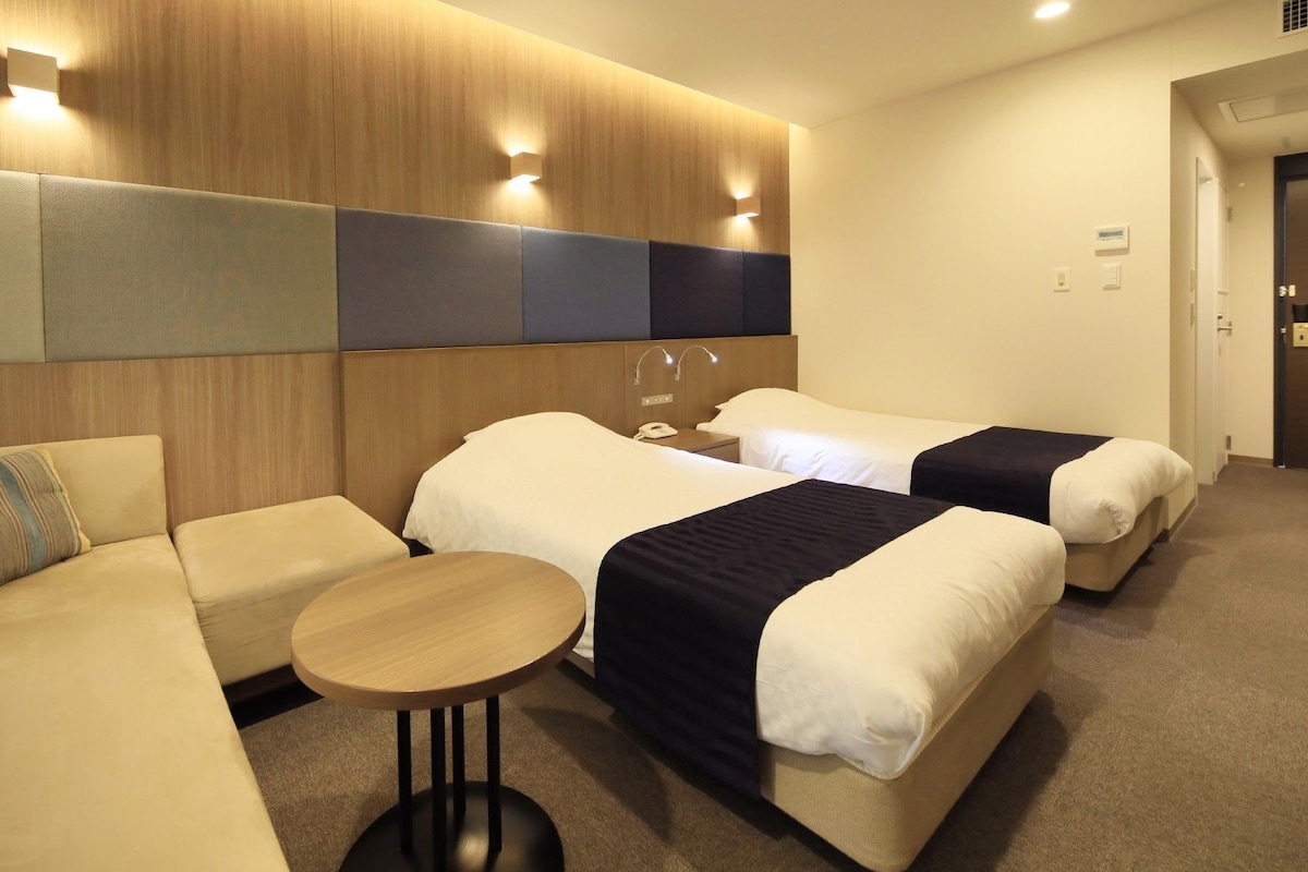 A hotel room with a sofa, two single beds, a small table, and a wooden headboard.