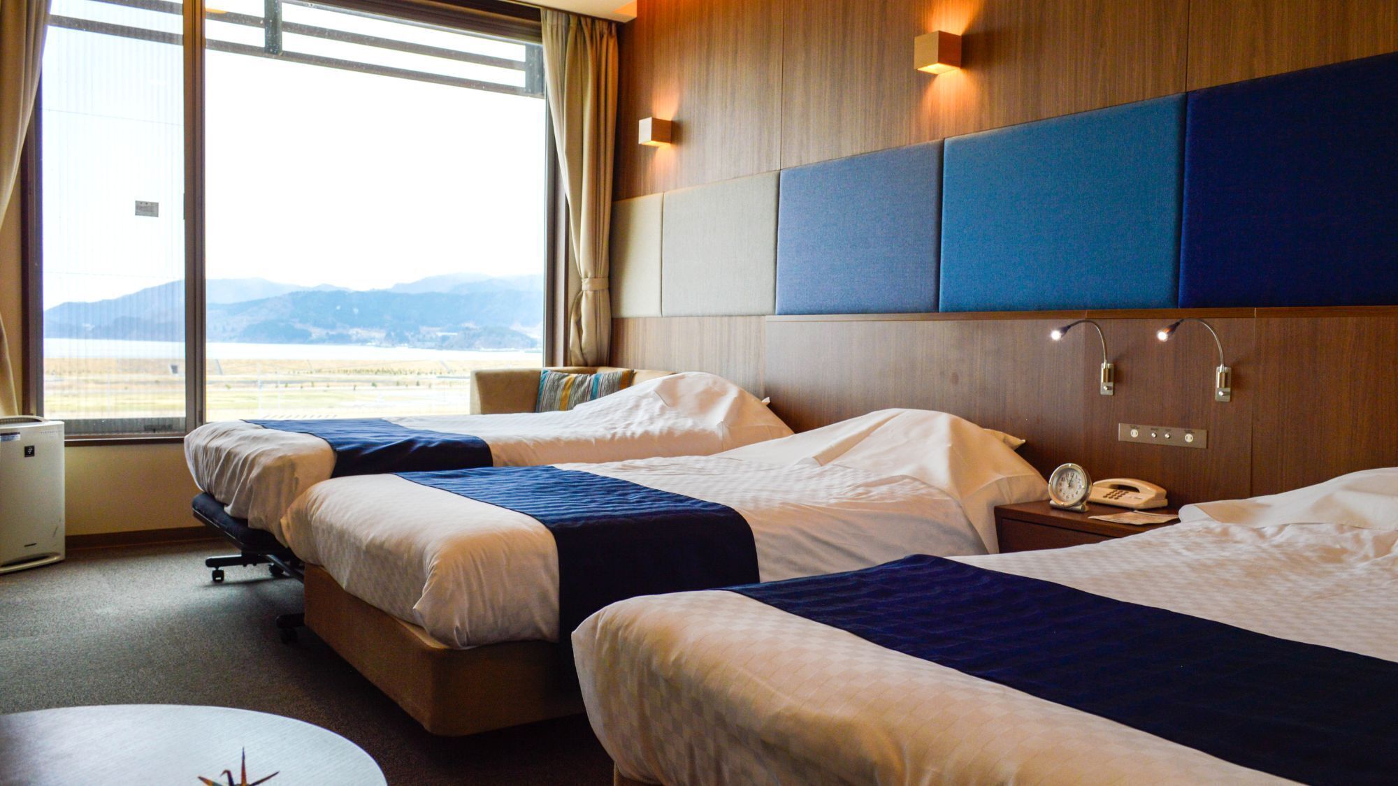 A hotel room with two beds, a desk, a chair, a bedside table with a lamp, and a window with a view of the outdoors.