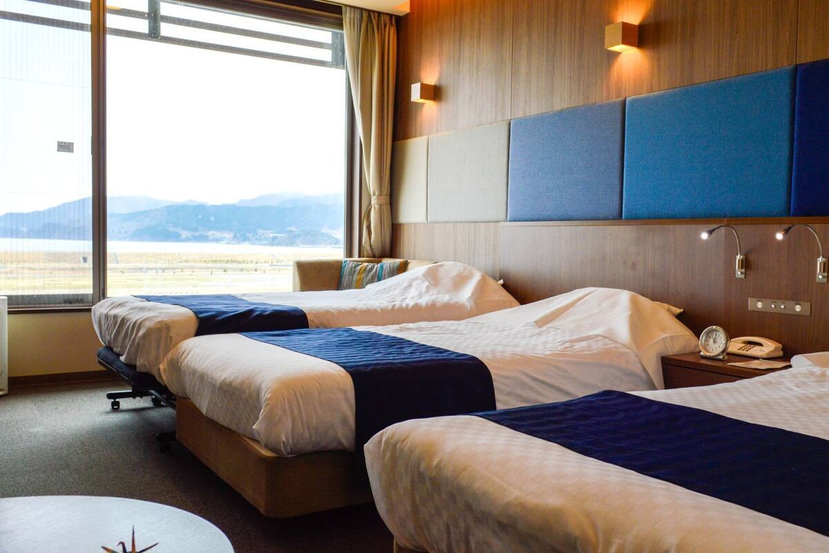 Ein Hotelzimmer mit zwei Betten, einem Schreibtisch, einem Stuhl, einem Nachttisch mit Lampe und einem Fenster mit Blick ins Freie.