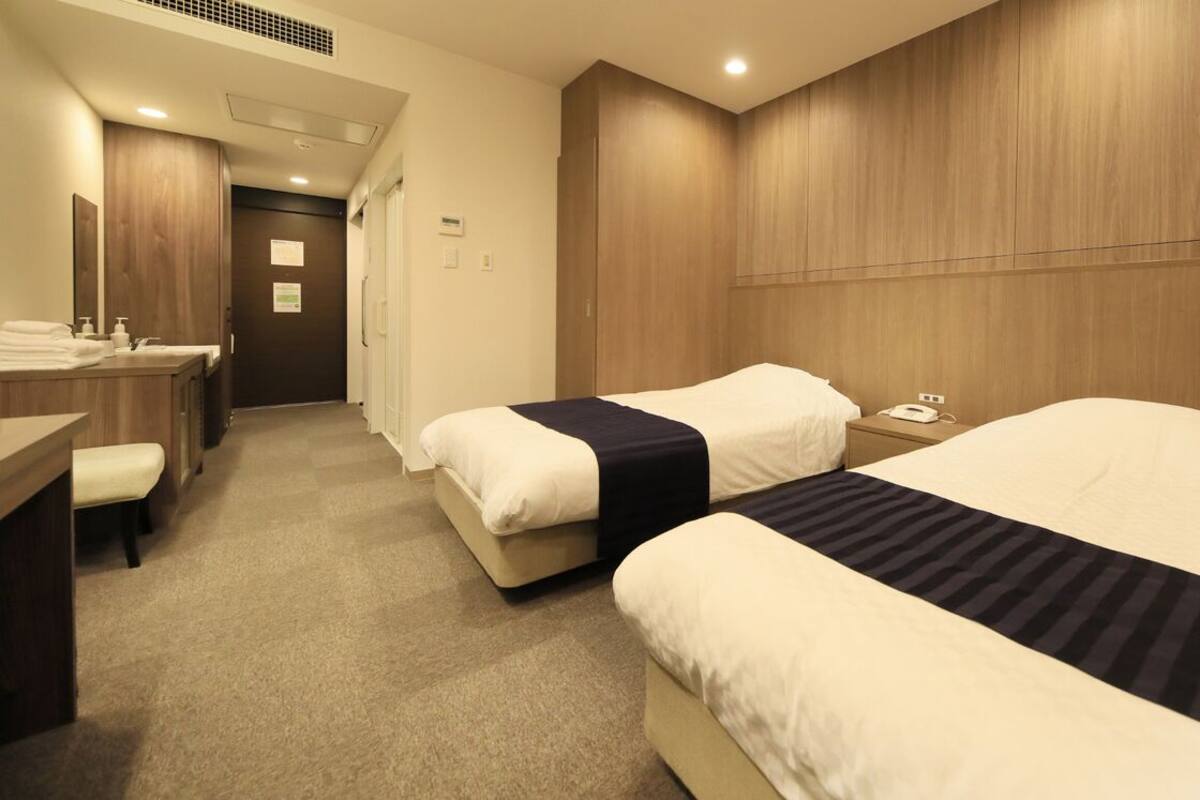 Ein Hotelzimmer mit zwei Betten, einem Schreibtisch, einem Stuhl, einem Spiegel und einem Holzkopfteil.