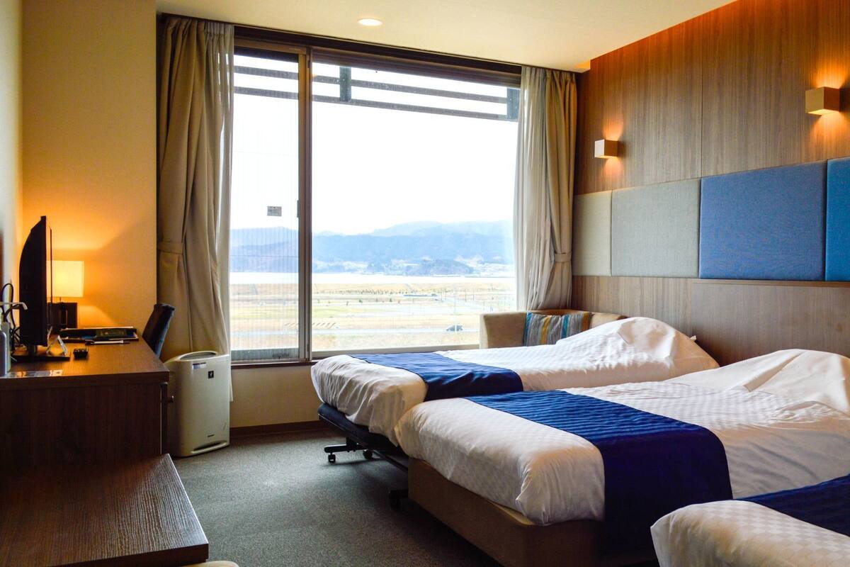 Ein Hotelzimmer mit zwei Betten, einem Schreibtisch mit Computer, einer Lampe und Blick auf das Flughafengelände.