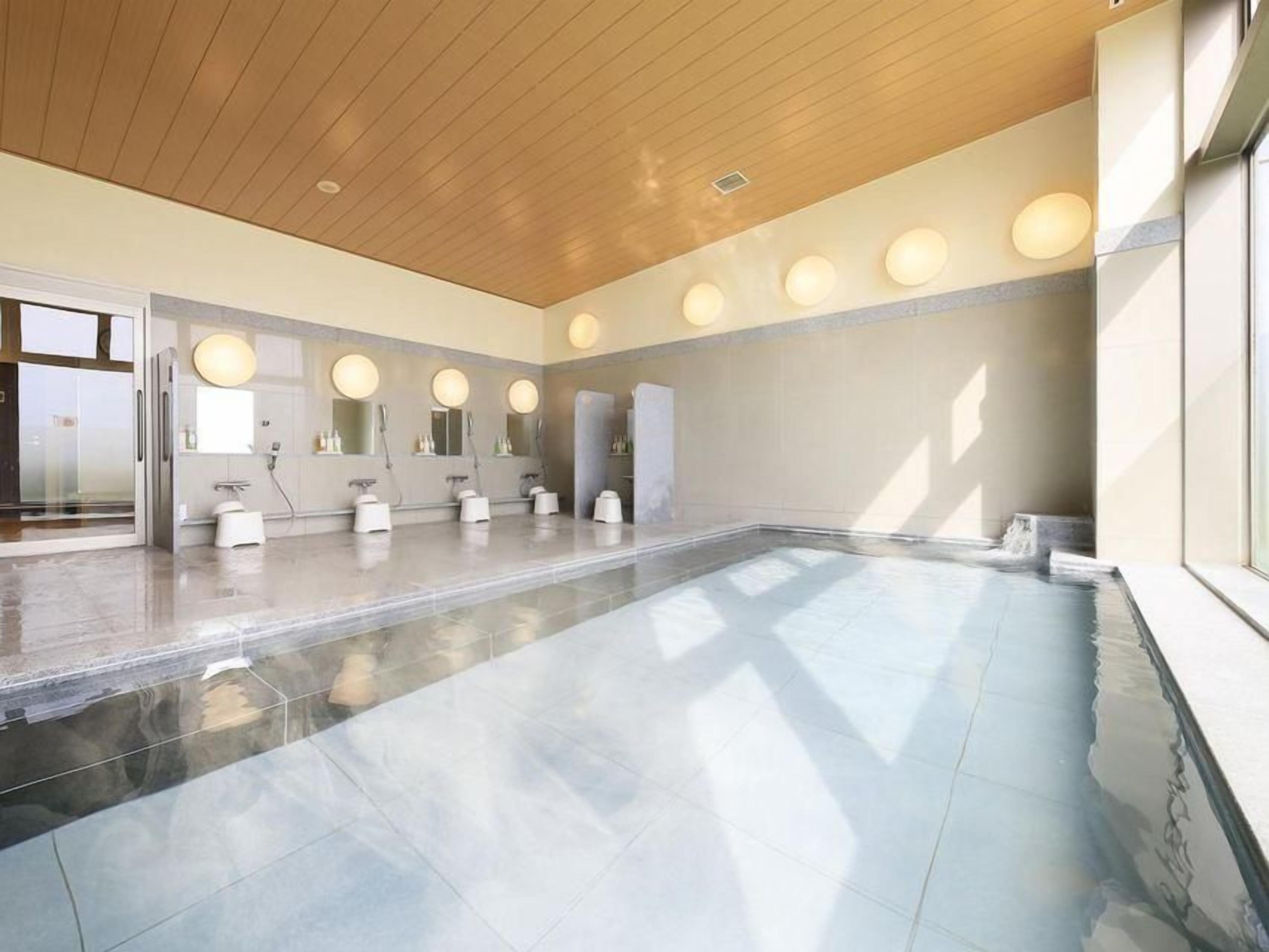 A modern indoor swimming pool with multiple lanes, a wooden ceiling, and round mirrors above the sinks.