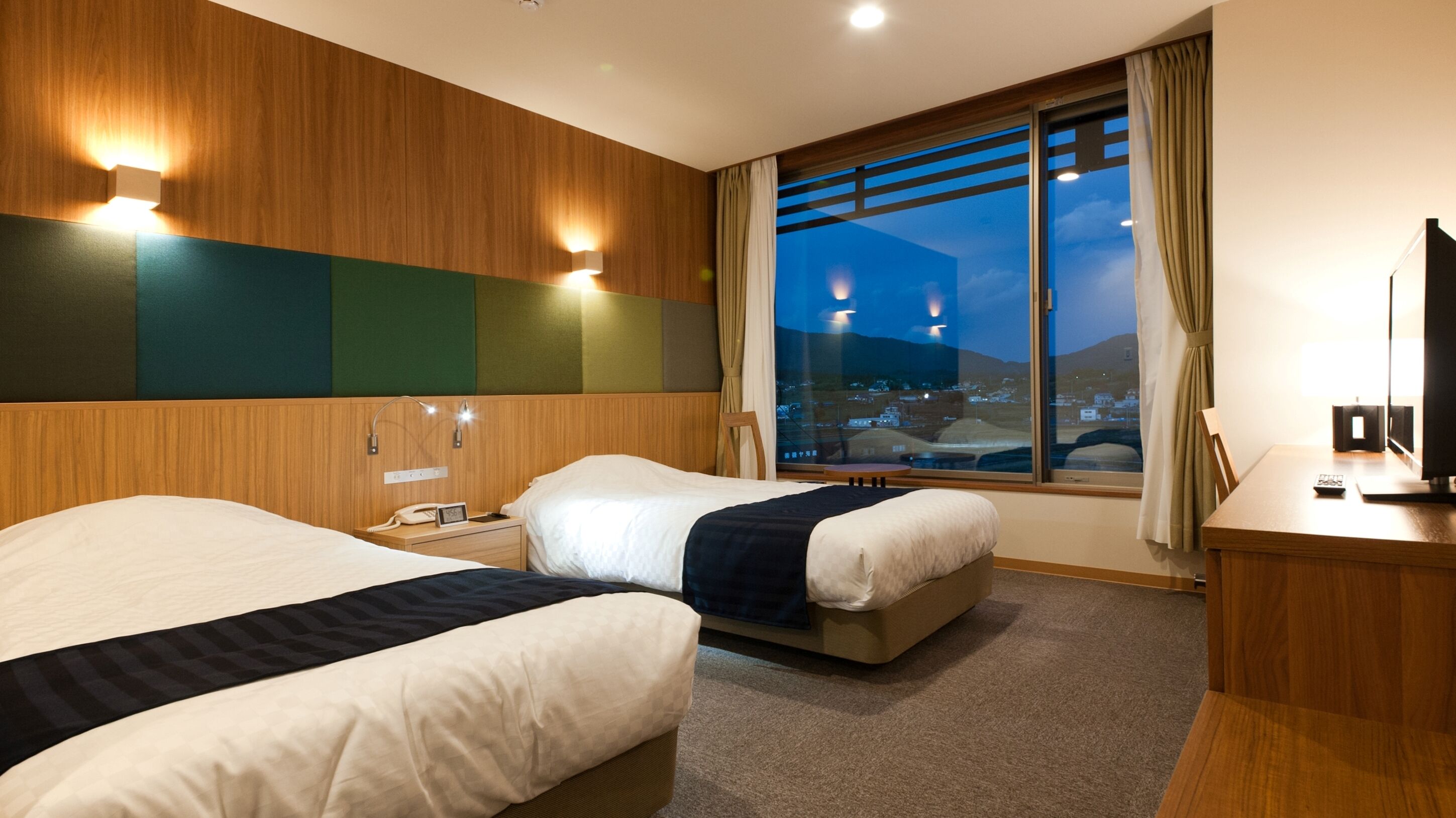 A hotel room with two beds, a desk, a chair, a TV, and a window with a city view.