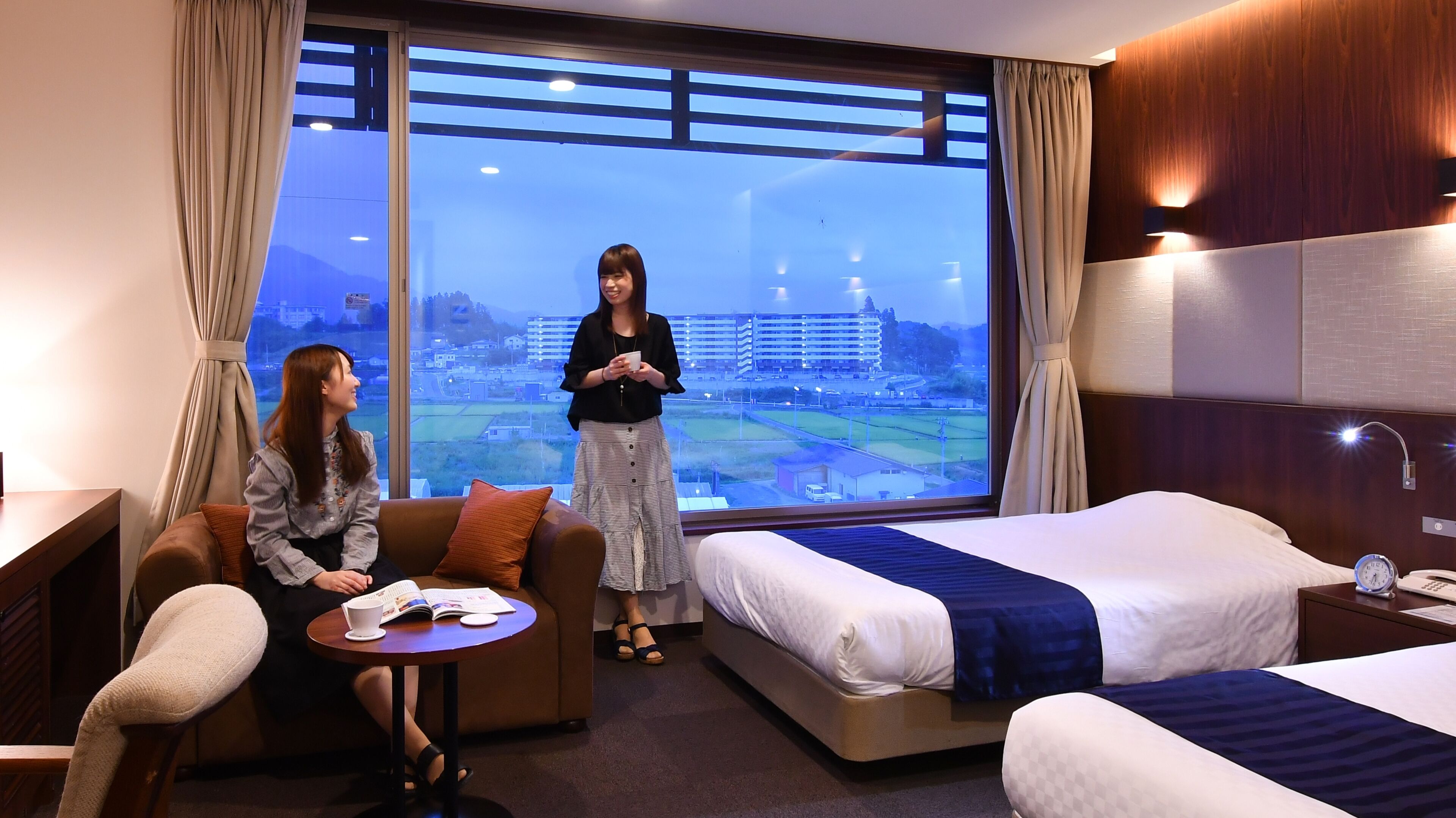 A hotel room with a large window, a sofa, a bed, and a small table with a book and a cup.
