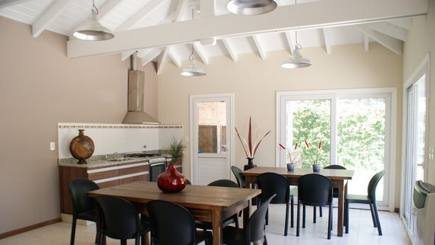 A dining area with a wooden table and chairs, a kitchen in the background, and a large window with a view of greenery.