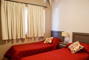 A room with two beds, each with a red bedspread and floral pillows, a nightstand with a lamp, and a window with curtains. - Descanso del Molino (Luján)