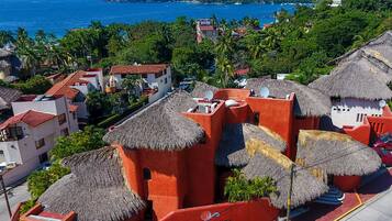 A coastal hotel with thatched-roof buildings, a paved road, and a view of the sea and hills in the background.