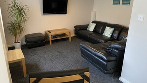 A living room with a black leather sofa, a wooden coffee table, a television on the wall, a potted plant, and a small table with a chair. - Ivy Cottage Serviced Accommodation (Aberdeen)