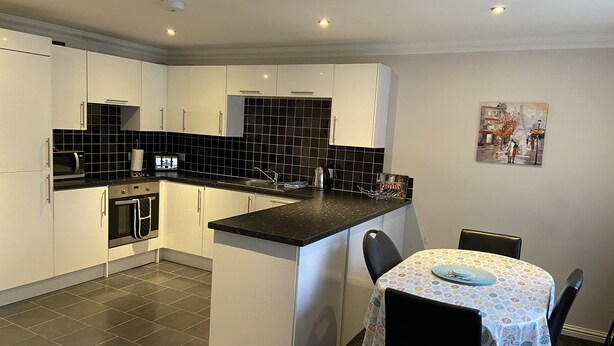 A modern kitchen with white cabinets, a black tiled backsplash, and a small dining area with a table and chairs. - Ivy Cottage Serviced Accommodation (Aberdeen)