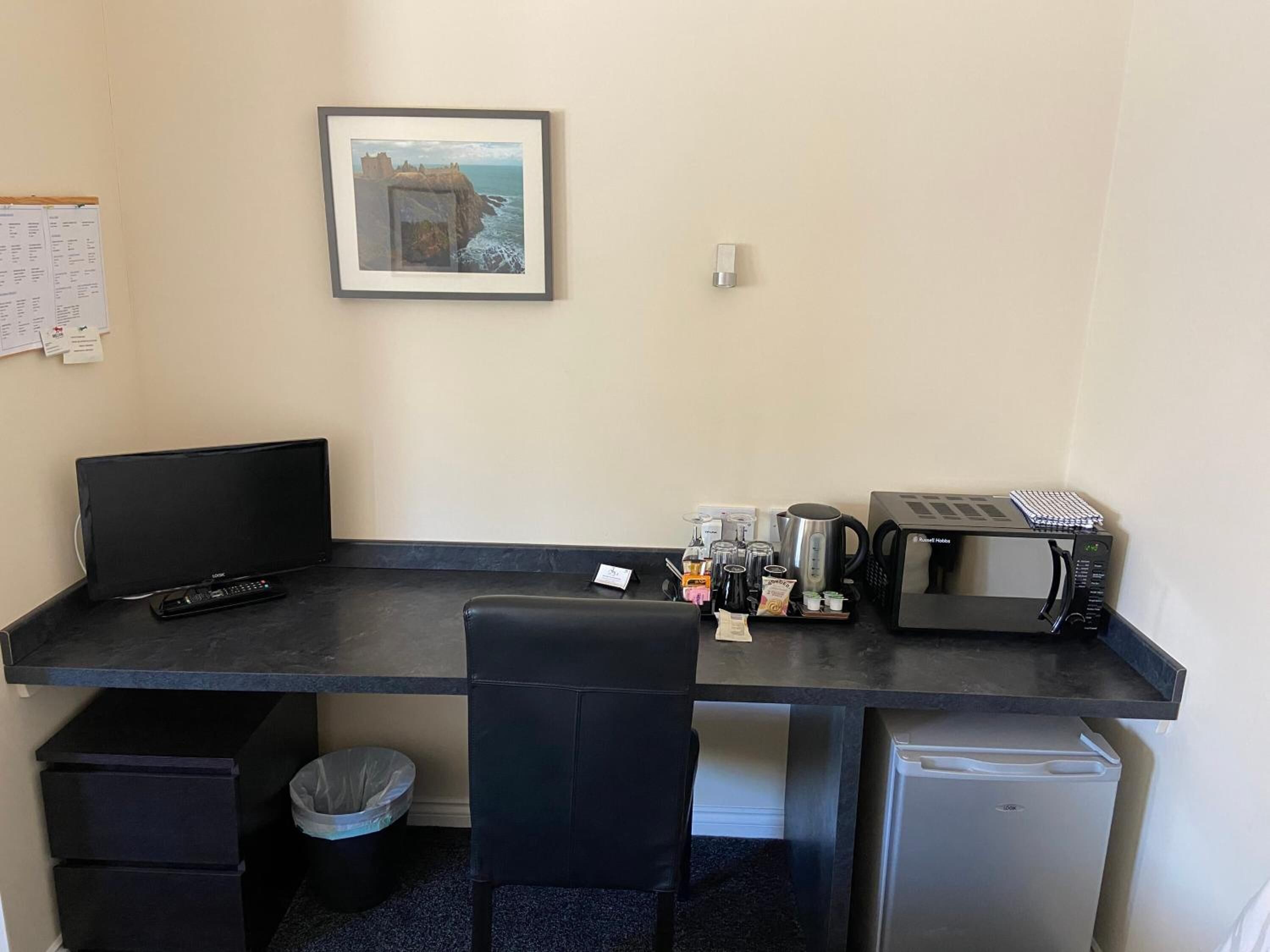 A compact hotel room with a desk, a computer, a small refrigerator, a microwave, a coffee maker, and a trash bin.