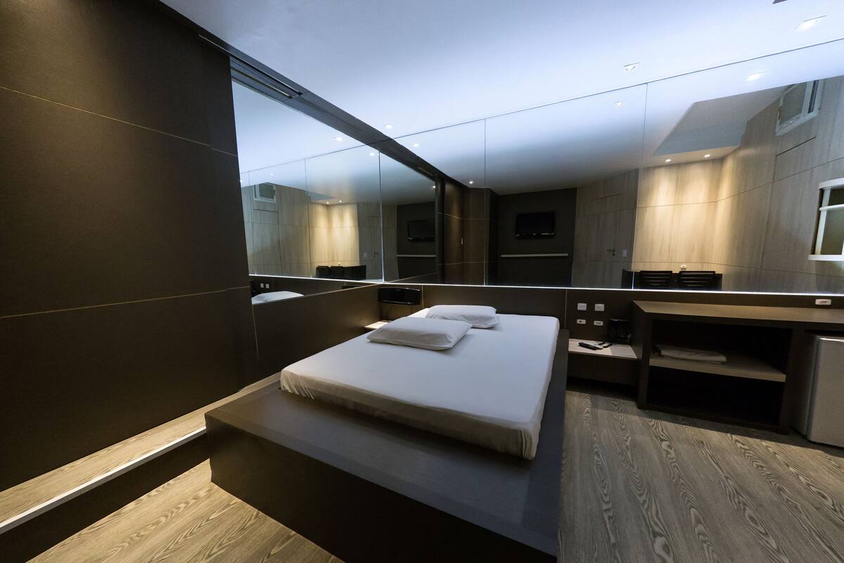 Una habitación de hotel moderna con una cama grande, un escritorio empotrado, un espejo y una pequeña cocina.