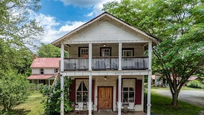 Uma casa de dois andares com varanda, um alpendre com assentos e uma entrada com calçamento de brita.