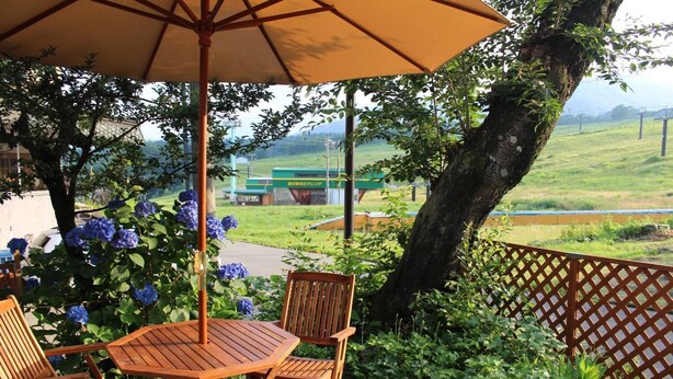 Une terrasse extérieure avec une table et des chaises en bois, un grand parasol et une vue sur un paysage verdoyant, avec un bâtiment en arrière-plan.