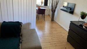 Ein schmaler Flur mit Holzboden, einer dunklen Couch, einem kleinen Tisch mit Pflanze und einem Spiegel an der Wand.