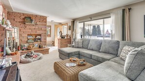 Un salon chaleureux avec une cheminée en briques, un canapé d’angle, un pouf en osier et une grande fenêtre agrémentée de rideaux.