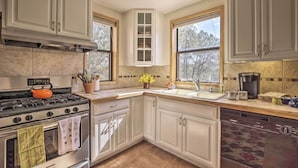 Una cocina con armarios blancos, estufa, horno y lavavajillas. Hay ventanas, un fregadero y una encimera con diversos objetos.