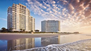 Ein Strandhotel mit mehreren Etagen, ein sandiger Strand im Vordergrund und ein klarer Himmel mit Wolken.