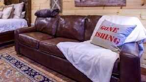 A wooden cabin interior with a brown leather couch, a bed with a wooden headboard, and a painting of horses on the wall.