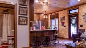 A rustic cabin interior with a wooden ceiling, a kitchen area with bar stools, and a cozy seating area with a fireplace.