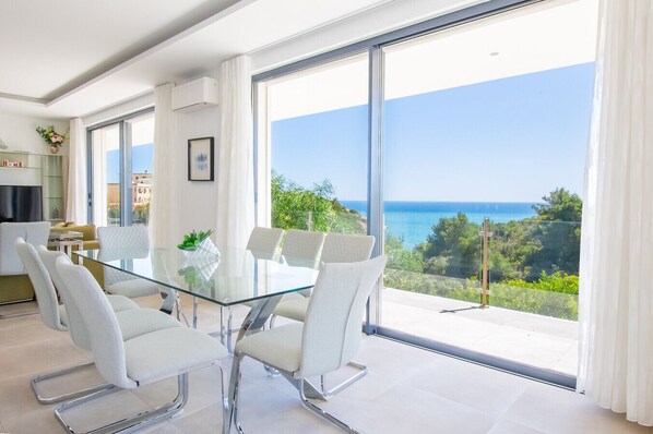 Une pièce moderne avec une table et des chaises en verre, de grandes fenêtres donnant sur la mer et un coin cuisine.