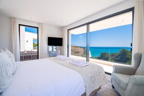 Ein Hotelzimmer mit einem großen Bett, einem Fernseher, einem Sessel und einem Balkon mit Meerblick.