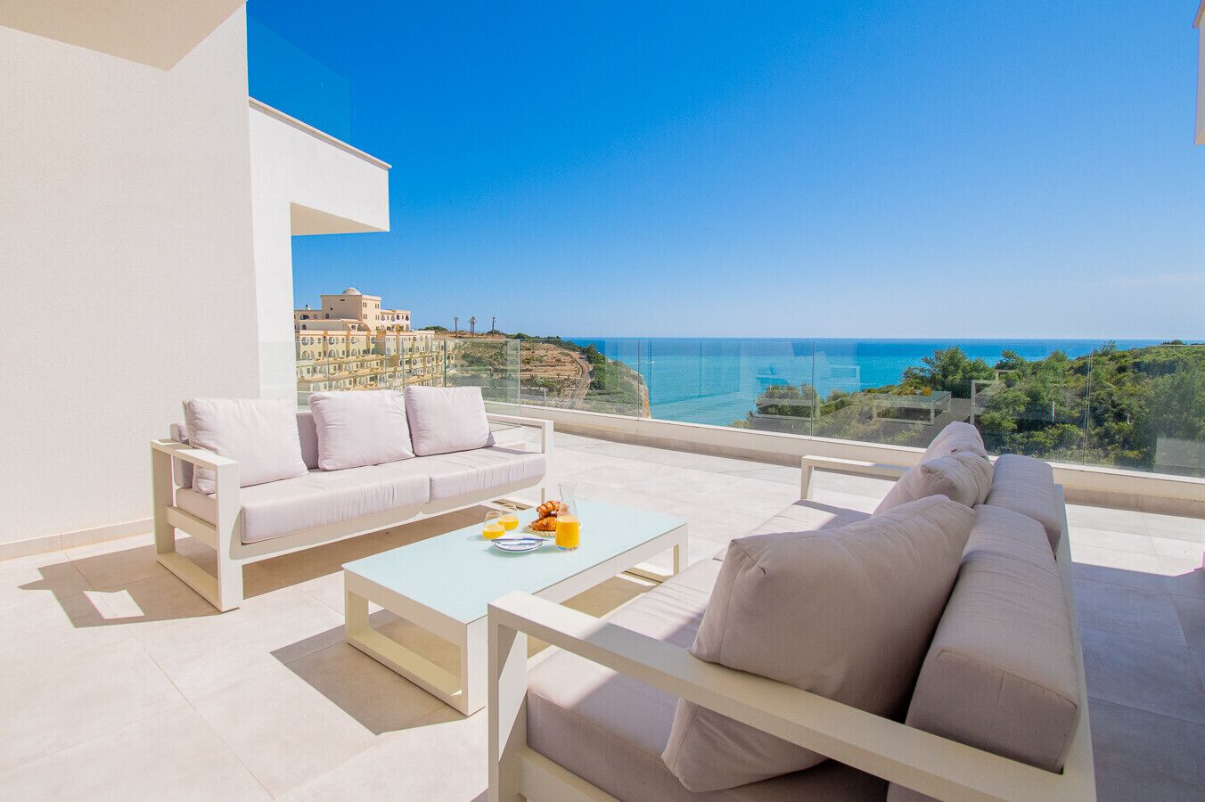 A modern outdoor seating area with a sofa and chairs, a coffee table, and a view of the sea and buildings.
