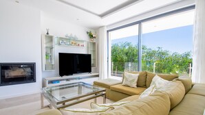A modern living room with a flat-screen TV, a glass coffee table, a beige sofa, and a large window offering a view of greenery. - Villa Centeanes Luxury villa,enviable position stunning sea views! (Carvoeiro)