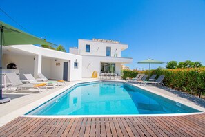 Un'area piscina moderna con lettini, un pontile in legno e un edificio bianco con tetto piatto.