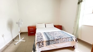 A bedroom with a bed, a nightstand, a bedside table, a fan, and a window with curtains.