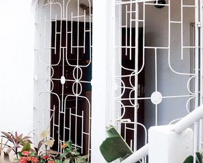 Una reja metálica blanca con un diseño geométrico, una pared blanca y una planta con flores rojas.