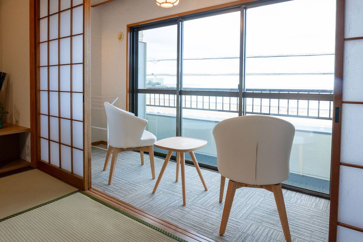 A traditional Japanese room with tatami flooring, a sliding shoji screen, a television, and two white chairs by a large window.