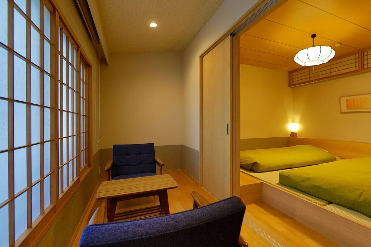 A traditional Japanese-style room with tatami flooring, a low table, a chair, and a bed with green bedding.