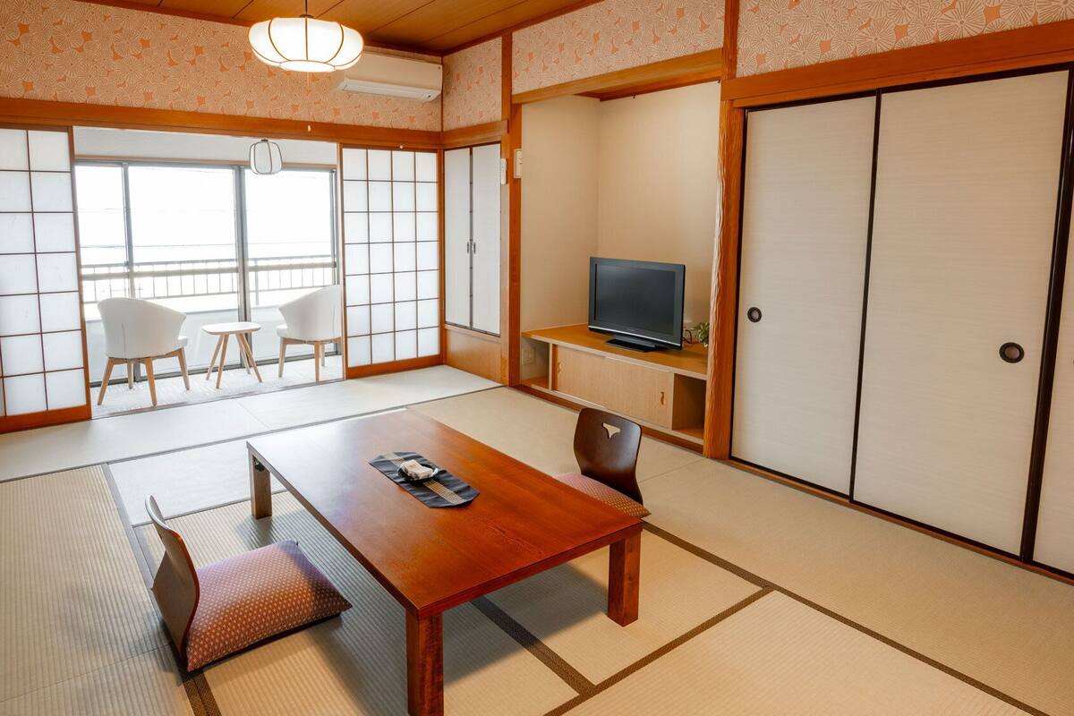 A traditional Japanese room with tatami flooring, a low wooden table, a television on a shelf, and sliding doors.