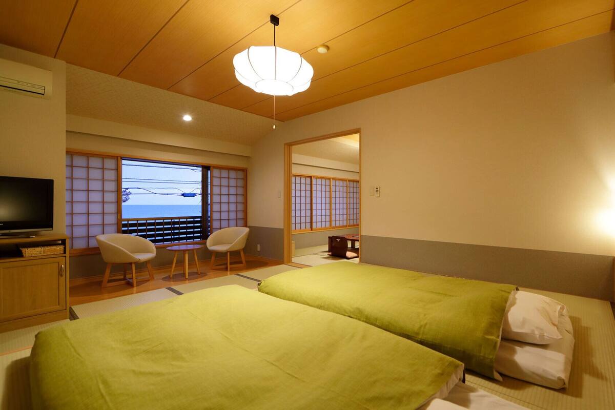 A hotel room with two beds, a TV, a seating area with chairs, and a view of the sea.